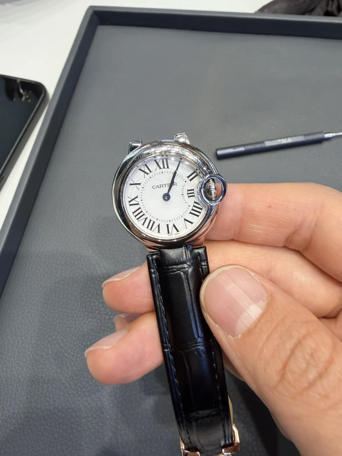 Front of a Cartier Ballon Bleu 28mm with a white dial, Roman numerals, and a black leather strap, held in a hand